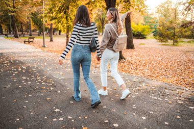 Two friends walk through a park covered in autumn leaves, laughing and enjoying the crisp air on a sunny day.