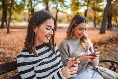 Two girls sit on a bench covered in golden leaves, happily texting on their phones amidst the vibrant colors of autumn.