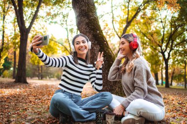 Two girls sit on the ground among fallen leaves, enjoying a lively moment and capturing memories with their phone while surrounded by autumn colors.