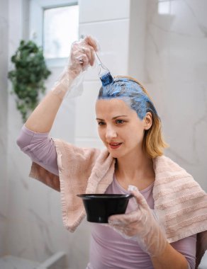 Smiling woman applying blue dye on her hair, sitting in the bathroom