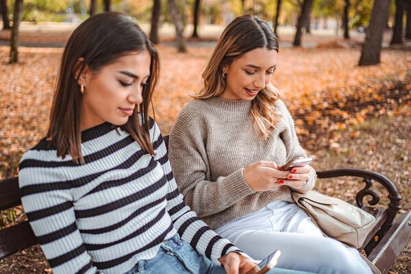 Two girls sit on a bench in a park filled with colorful autumn leaves, chatting and enjoying their phones while surrounded by natures beauty.