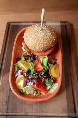 Canlı bir düzenleme, rengarenk sebze ve dilimlenmiş domates karışımının yanında tahtadan bir tepside sunulan susamlı çörek hamburgerini içerir..