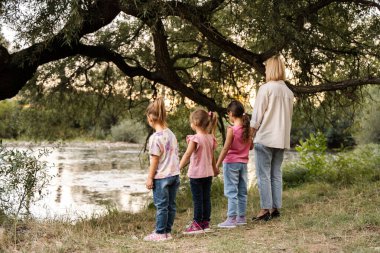Mother with three daughters holding hands looking at river under tree in late afternoon
