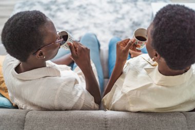 Two black women enjoying a relaxing moment on the sofa, sipping coffee in their cozy living room
