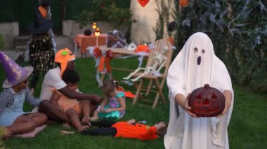 Boy in a ghost costume holding a jack-o-lantern scaring a group of children at a Halloween party