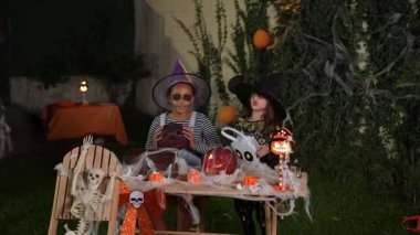 Two happy girls in witch costumes playing with halloween decorations in a garden at night