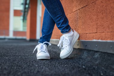 Deri beyaz Sneaker şehirde duvara eğilme ile kadın, kot ve spor ayakkabı giyen kadın bacaklar yakın yan görünüm. Moda konsepti