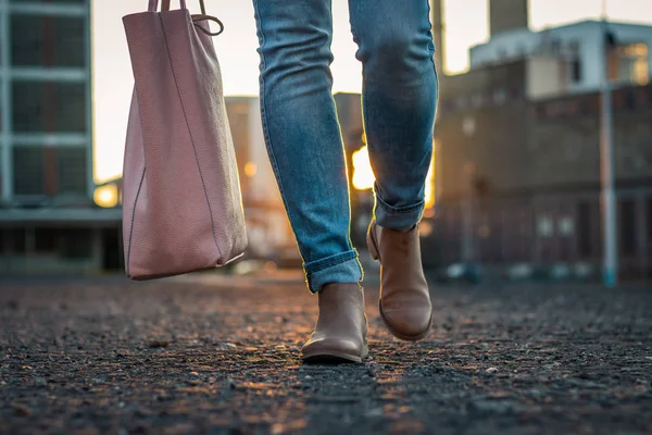 Woman in jeans and stylish leather shoe with bag walking in city during sunset. Casual clothing in city