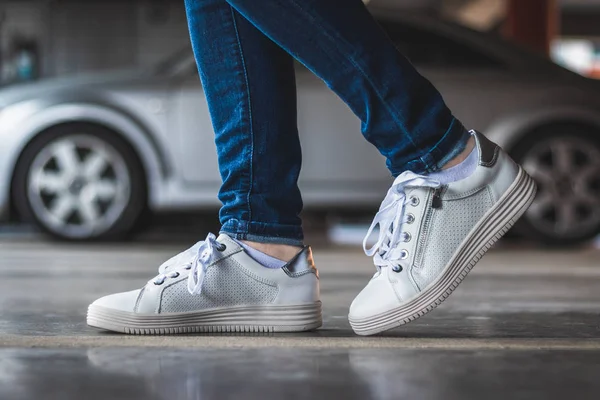 Woman with white sneakers standing in parking lot, blurry car in background. Legs with unbranded sport leather shoes