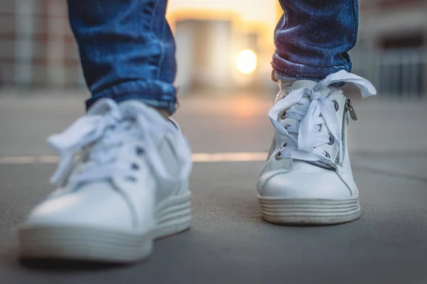 Şehirde gün batımında beyaz spor ayakkabıları var. Sokakta spor ayakkabısıyla yürümek. Açık havada ayakkabılara yakın ön görünüm. 