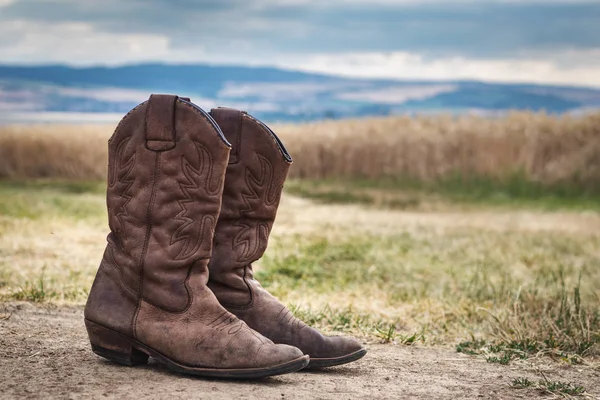 牛仔靴在农村的场景与喜怒无常的天空 户外的旧棕色皮靴 狂野的西部复古风格 — 图库照片