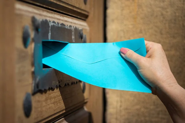 Woman inserting blue envelope into mail slot. Delivering letter by mail carrier. Close-up hand with envelope