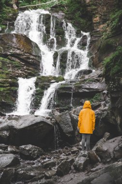 sarı yağmurluk ayakta ve şelale arayan bir kadın. Hiking kavramı