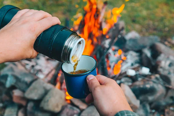 man pour tea in metal cup. camp fire on background. hiking