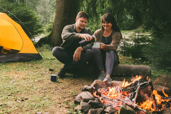couple sitting near fire and drink hot tea. hiking concept. car with tent on background