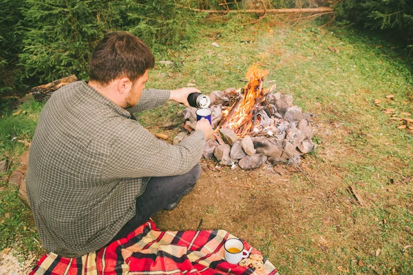 man pouring tea in metal cap near camp fire. hiking concept