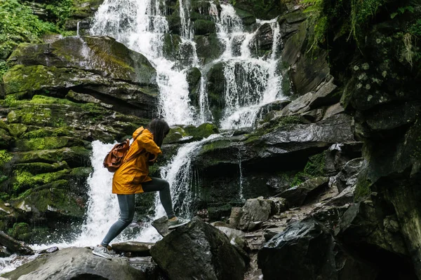 woman jump from rock to rock in forest in yellow raincoat. water fall on background