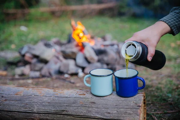 hand pour tea into metal cups. camp fire on background. hiking concept