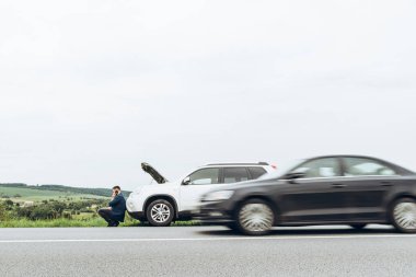 kırık arabanın yakınında telefon ile oturan adam yardım arıyor. Road trip sorunları