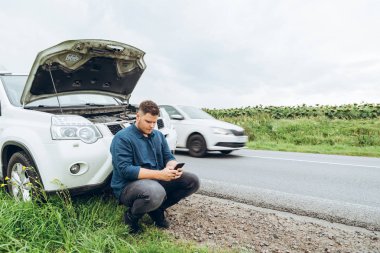 kırık arabanın yakınında telefon ile oturan adam yardım arıyor. Road trip sorunları