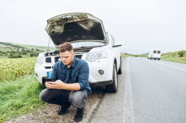 kırık arabanın yakınında telefon ile oturan adam yardım arıyor. Road trip sorunları