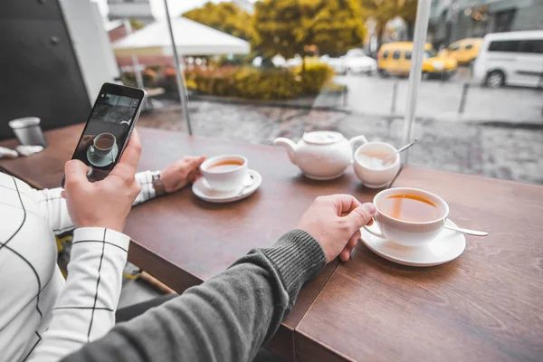 寒い秋の日に暖かいお茶を飲むカフェに座っているカップル 女性は Instagram の電話で写真を撮る 最初人の眺め — ストック写真