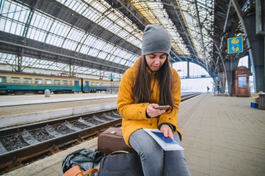 Genç kadın çantasını tren istasyonunda oturmak ve telefonunu kontrol edin. Tren bekliyor