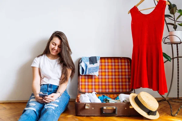 smiling woman sit on floor near valise with clothes. packing before trip. travel concept. copy space