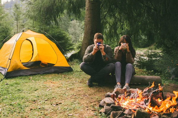 couple sitting near fire and drink hot tea. hiking concept. tent on background