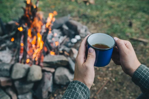 first point of view. man drinking tea in metal cup near camp fire. hiking