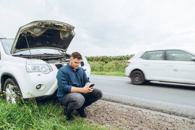 kırık arabanın yakınında telefon ile oturan adam yardım arıyor. Road trip sorunları