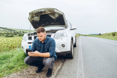 kırık arabanın yakınında telefon ile oturan adam yardım arıyor. Road trip sorunları