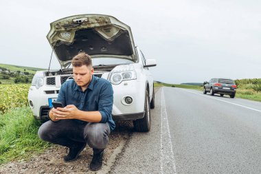 kırık arabanın yakınında telefon ile oturan adam yardım arıyor. Road trip sorunları