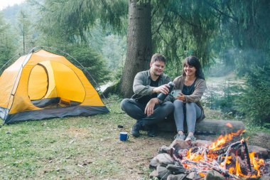 Yangın ve içecek sıcak çay oturan bir çift. Hiking kavramı. araba arka plan üzerinde çadır ile