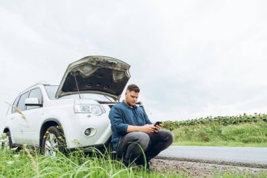 kırık arabanın yakınında telefon ile oturan adam yardım arıyor. Road trip sorunları