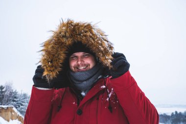 kırmızı kış ceket kürk başlık arka plan üzerinde kar alan genç adam gülümseyen.