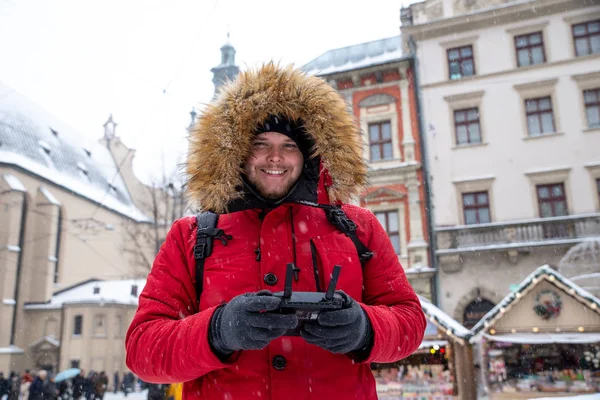 Retrato de hombre adulto joven en la capucha con piel en la plaza de la ciudad de invierno con controlador de drones en las manos — Foto de Stock