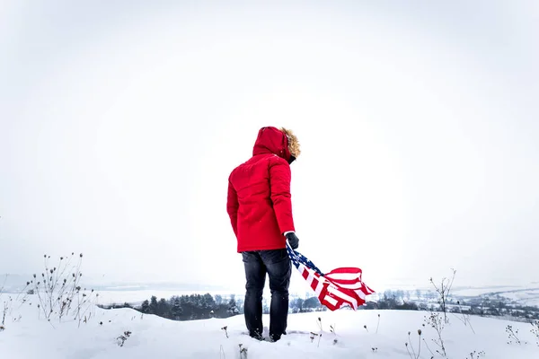 Man in red winter coat holding usa flag in hand on wind in the middle of the snowed field — Stock Photo, Image