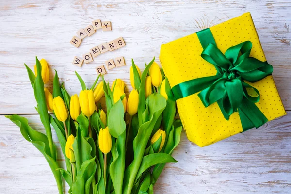 happy woman\'s day text. tulips overhead on white wooden background with yellow gift box