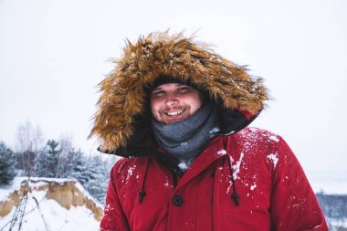 kırmızı kış ceket kürk başlık arka plan üzerinde kar alan genç adam gülümseyen.