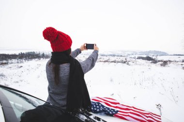 ABD bayrağı alarak resim telefonunda ile Cipin kaputunun üzerinde oturan genç yetişkin kadın gezgin. araba seyahat