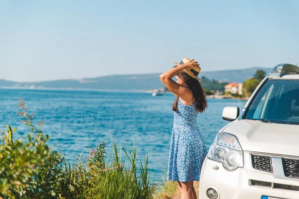 妇女在蓝色礼服站立在白色汽车附近在海边与美丽的看法在海湾和山 夏季旅游理念 — 图库照片
