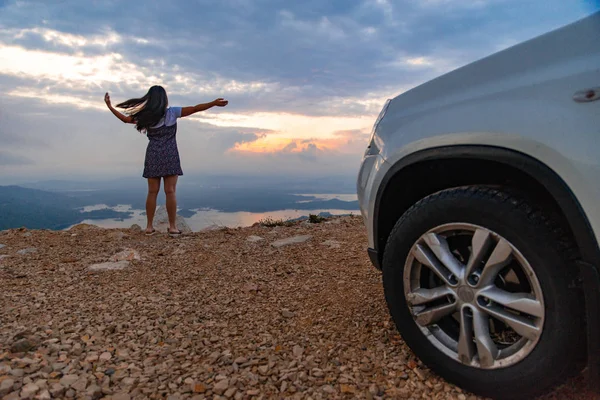 woman standing on cliff near car with beautiful view of lake on sunrise. road trip