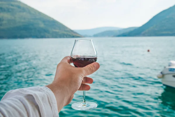 男の手は海と山を背景に赤ワインのガラスを保持しています ライフ スタイル — ストック写真