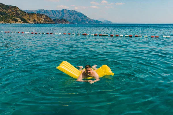 Mladý dospělý muž plavání na žlutou nafukovací matraci v modrém moři. — Stock fotografie