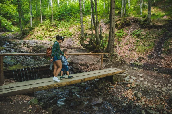 Mama cu copil traversând râul prin pod în pădure — Fotografie, imagine de stoc