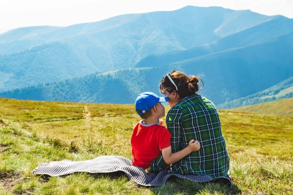 Madre joven con un niño sentado en una manta en la cima de la montaña. hermosa vista del paisaje en el fondo —  Fotos de Stock