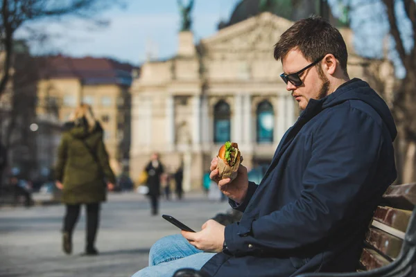 Homem comendo hambúrguer surf internet no telefone sentado no banco — Fotografia de Stock