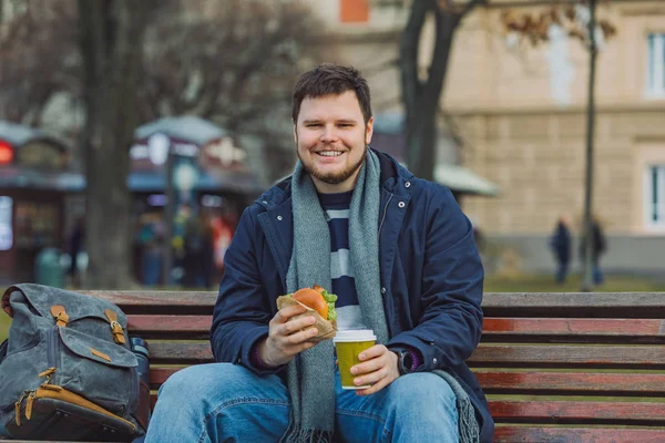 Jonge man eten hamburger en drinken koffie zittend op stad bankje glimlachen — Stockfoto
