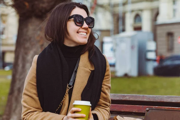 Young stylish smiling woman in sunglasses drink coffee to go while sitting on bench — Stock Photo, Image